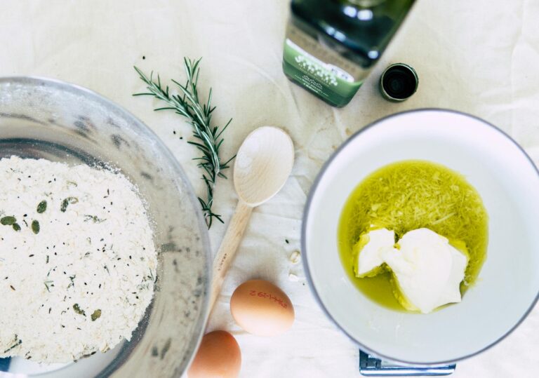 Flat lay of baking ingredients including flour, eggs, and olive oil with rosemary on wooden table.