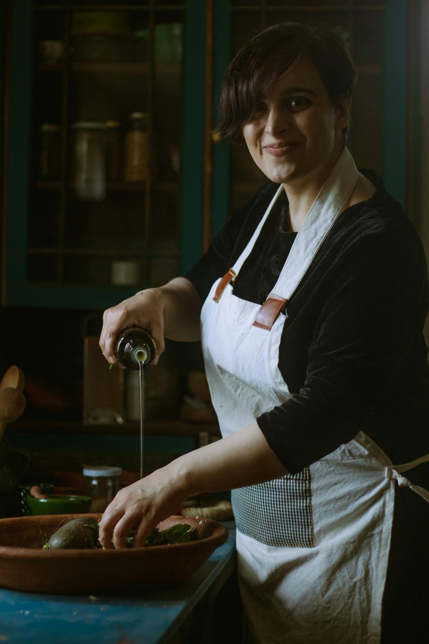 Woman in a cozy kitchen pouring olive oil onto vegetables while smiling.